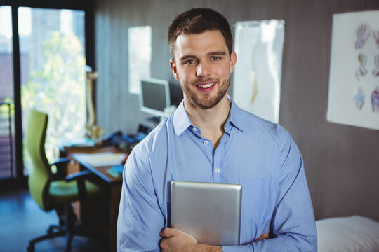 Man in his 20s standing in clinic office holding tablet beside spine model and anatomical charts