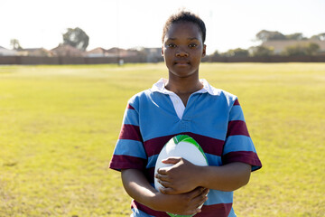 African American teenage girl holding rugby ball wearing striped jersey on suburban sports field