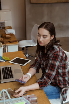 Woman wearing red plaid shirt examining credit card while sitting at office desk with laptop