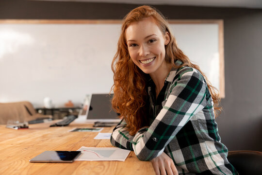 Red-haired woman leaning forward arms resting on table with tablet in meeting room, copy space