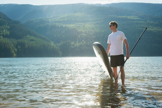 Fototapeta Person standing in calm lake holding paddleboard and paddle, forested hills reflecting sunlight