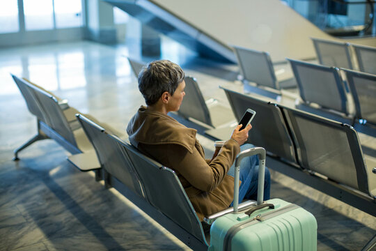 Traveler sitting on bench holding smartphone and coffee with mint-green suitcase in airport lounge
