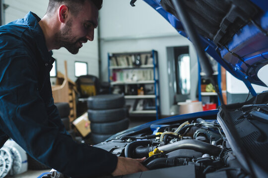 Male mechanic wearing coveralls examining engine under open hood with tires at garage, copy space