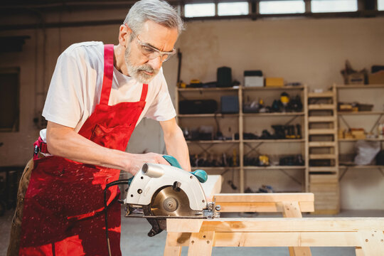 Man operating circular saw on wooden workbench in woodworking workshop with safety glasses
