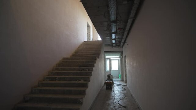 Corridor stretches toward stairwell leading upstairs. Exposed concrete and dusty surfaces show early progress. Empty rooms echo with steps