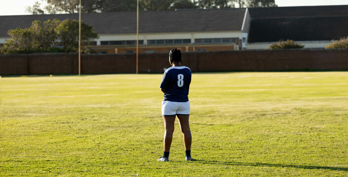 Athlete wearing blue jersey standing with arms folded on grass field near goalposts, under sunlight