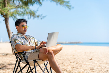 Asian man remote working corporate business on laptop computer while travel the sea on summer beach holiday vacation. Workation, work from anywhere with wireless technology and digital gadget device.
