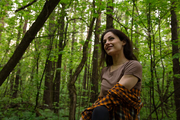 Naklejka premium Beautiful smiling happy woman hiking in a green forest relax sitting on the logs looking up on sunny spring weather. Girl rest enjoy, breathing deep in style fashion clothes among the woods