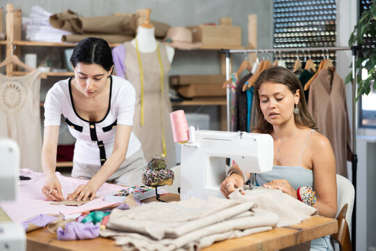 Female dressmakers sew clothes in the workshop. Seamstress works at the machine next to the fashion designer, who makes markings on the fabric - Powered by Adobe