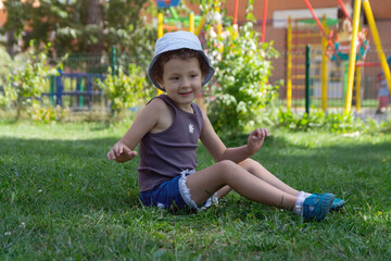 A child in a panama hat sits on the lawn near the playground