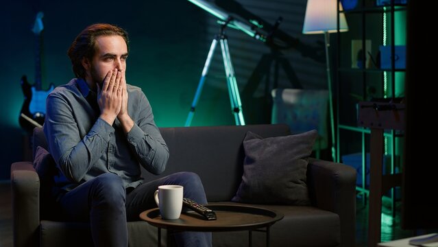 Man at home shocked by thriller film on TV, drinking coffee. Stupefied person at home left with eyes wide open by dramatic plot twist, watching suspenseful movie on TV, enjoying beverage, camera A
