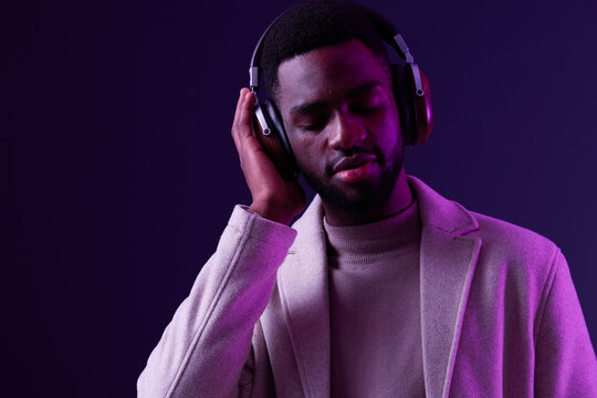 A young Black man enjoying music with headphones, dressed in a stylish beige coat and light sweater, set against a vibrant purple background, conveying relaxation and contentment