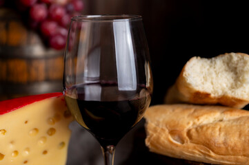 Glass of red wine with a bottle serving on a wooden table with cheese, bread, and salami. Fresh grapes, a knife, and a board. Still life photo.