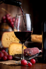 Glass of red wine with a bottle serving on a wooden table with cheese, bread, and salami. Fresh grapes, a knife, and a board. Still life photo.