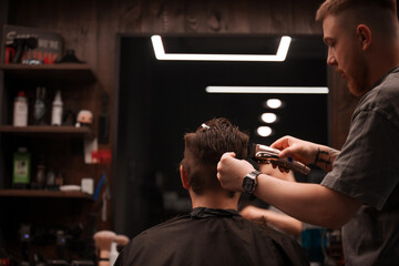 Barber uses electric clippers to trim the back of a client's head in a modern barbershop, seen from behind. Shelves with products and a lit mirror frame the stylish workspace.