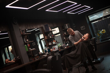 A young barber with a trimmed beard carefully shapes a client’s beard in a modern barbershop with stylish lighting and mirrors, capturing a moment of precision and grooming care..