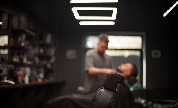 Fototapeta Modern barbershop interior with a black barber chair in focus, while a barber grooms a male client in the blurred background under geometric LED ceiling lights. Contemporary grooming scene..