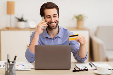 Obraz premium Cheerful Man Calling To Bank By Phone, Holding Credit Card Sitting At Laptop In Modern Office. Banking Assistance Service, Online Shopping Concept. Selective Focus