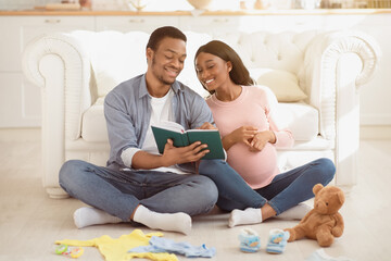 Joyful black couple making checklist of baby things for maternity hospital, writing in notebook at home. Positive future parents preparing for childbirth, packing stuff for clinic