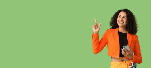Stylish young African-American woman with mobile phone pointing at something on green background with space for text