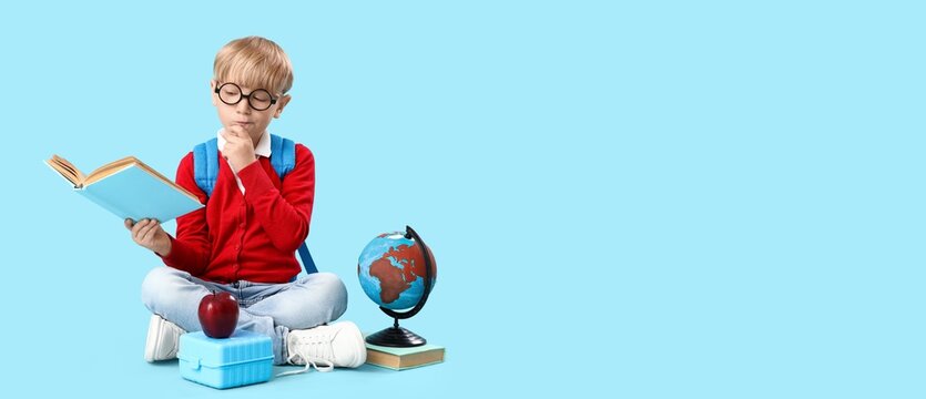 Little thoughtful schoolboy with lunchbox, apple and globe reading book on blue background with space for text