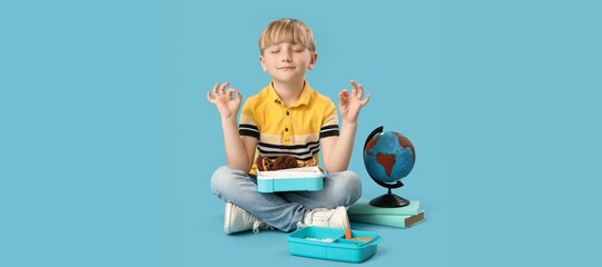 Little boy with lunchboxes, books and globe meditating on blue background