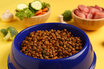 Bowls with dry pet food, raw meat and natural products on yellow background, closeup