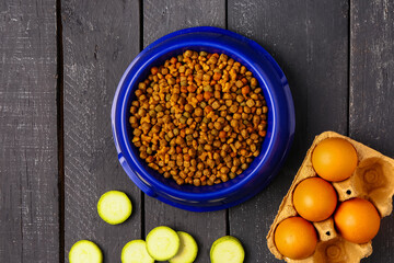 Bowl with dry pet food and natural products on dark wooden background