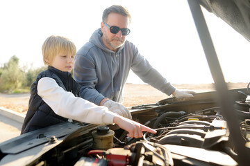 A middle-aged father and a preteen son work on a car during a road trip