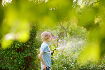 Funny little boy watering plants and playing with garden hose with sprinkler in sunny backyard. Happy child having fun with spray of water.