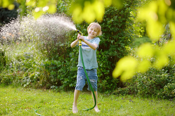 Funny preteen boy pouring cool water on himself and playing with garden hose with sprinkler in sunny backyard on hot summer day. Happy child having fun with water splashes.