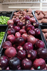 A bin overflowing with plump, dark red onions sits beside sweet potatoes in a grocery store