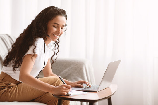 Young pretty brunette lady attending webinar from home, sitting on sofa, using laptop and headset, taking notes and smiling, enjoying online studying during COVID-19 pandemic, copy space