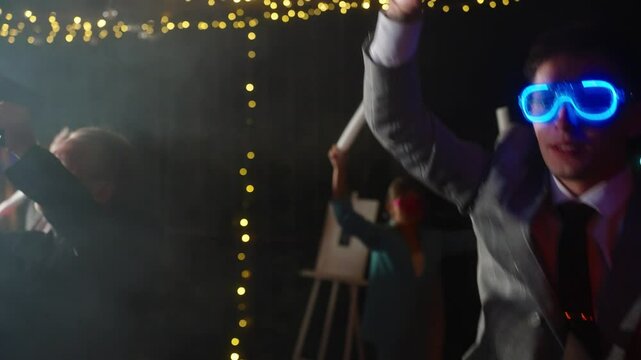 Close up of groom in a stylish suit dances joyfully at his wedding party, wearing glowing blue LED glasses. Concept of festive atmosphere, happy memories of celebrate college night party