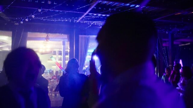 People dancing at a party with glowing LED sticks under colorful lights. Concept of celebrations wedding party, nightlife, event, many people dance, college party, bachelor party