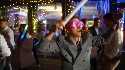 Stylish bride and groom wearing pink LED glasses dances enthusiastically at a wedding party, holding a glow stick. Concept of event, celebration, and nightlife themes, many people dance - Powered by Adobe