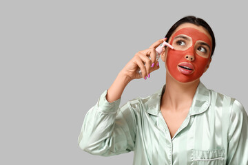 Beautiful young woman with facial mask and bottle of cosmetic product on grey background