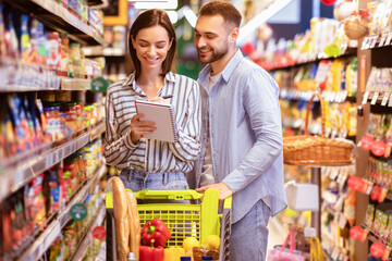 Portrait Of Two Happy Customers Holding Grocery Shopping Checklist, Writing In Notebook, Buying Products, Standing With Full Shop Cart In Supermarket. Young Millennial Family Choosing Healthy Food