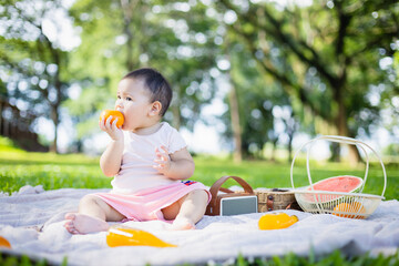 Baby enjoys picnic on blanket in park surrounded by green trees with fruit and basket nearby capturing family leisure moment with people concept in nature