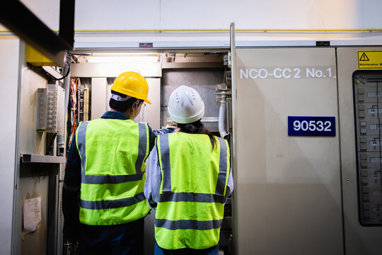Two engineers wearing insulated safety helmets and reflective vests inspect electric switchgear in gas insulated substation field ensuring electric grid operates safely and efficiently