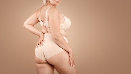 Young plus size woman in beige underwear posing over brown studio background, half-turned at...