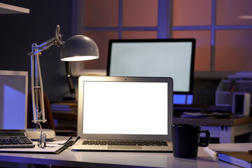 Blank laptop with glowing lamp on desk in dark office at night