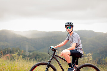 Young man riding a mountain bike outdoors on a cloudy day. He is wearing a helmet and sunglasses,...