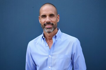 Smiling middle-aged man with close-cropped hair and beard wearing light blue button-up shirt against plain dark blue background