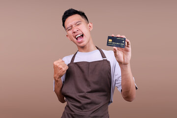 Excited Young Asian Man Wearing Brown Apron Holding Credit Card And Celebrating Victory Isolated On Beige Background