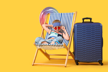 Cute little boy with inflatable ring and suitcase sitting in deck chair on yellow background. Travel concept