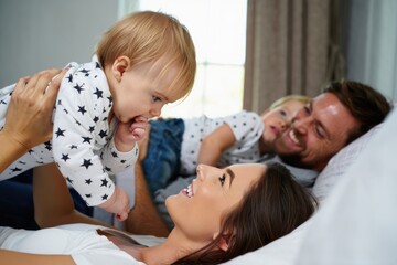 Obraz premium Happy family lying on bed with mother holding baby above her while father and young child smile in background in bright cozy room