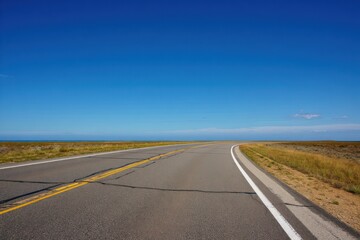 Fototapeta premium Empty curved road stretching through dry grassy landscape under clear blue sky with bright sunlight