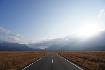 Long empty road stretching straight through dry golden fields towards distant mountains under a clear sky with bright sunlight breaking through clouds