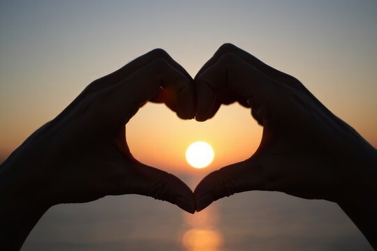 Silhouette of two hands forming a heart shape around the glowing sun during a peaceful sunset over calm water